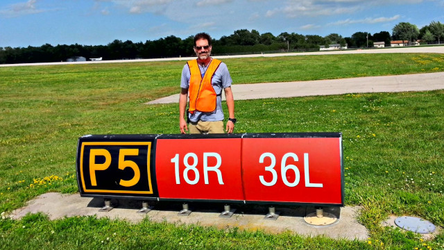 Ian stands in an orange safety vest near runway sign at Oshkosh in 2015